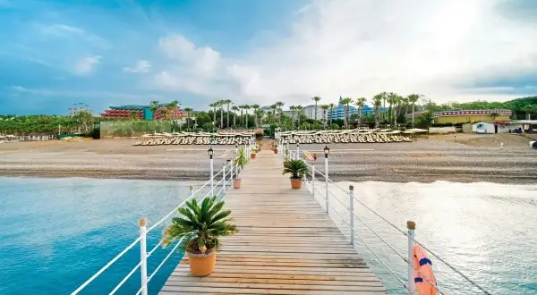 Drewniane molo prowadzi do plaży z rzędami leżaków i palm. W tle widać hotel, który zachęca do sprawdzenia mukarnas spa resort opinie.
