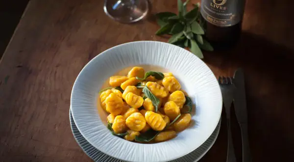 Gnocchis de potimarron aux feuilles de sauge, servis dans une assiette blanche sur une table en bois. Une bouteille de vin et un verre sont visibles en arrière-plan. Parfait pour une recette gnocchi potimarron.