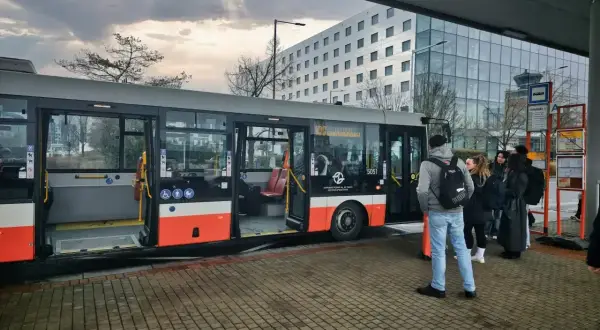 Ludzie czekają na autobus, który może być odpowiedzią na pytanie "jak dojechać do Pragi". W tle nowoczesny budynek i niebo.