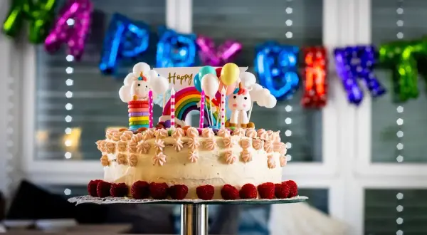Kolorowy tort z jednorożcami i tęczą, idealny na zabawy na urodziny 7 latka. Balony i napis "Happy Birthday" w tle.