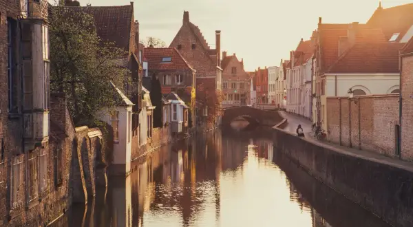 En kanal i Brygge, Belgien, med gamla tegelhus och en bro. Solen går ner och reflekteras i vattnet.