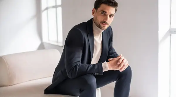 Mężczyzna w granatowym garniturze i beżowym golfie, prezentujący styl smart casual. Siedzi na kanapie, opierając dłonie na kolanach.