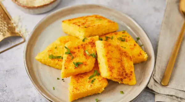 Gebratene Polenta-Ecken, italienisch garniert mit Petersilie, auf einem Teller. Daneben eine Reibe mit Parmesan.