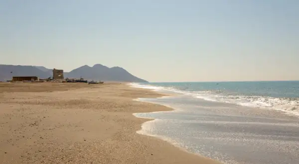 La playa de San Miguel, con sus aguas turquesas y arena dorada, se extiende hacia unas montañas. En la orilla, barcos y una torre antigua esperan la marea.