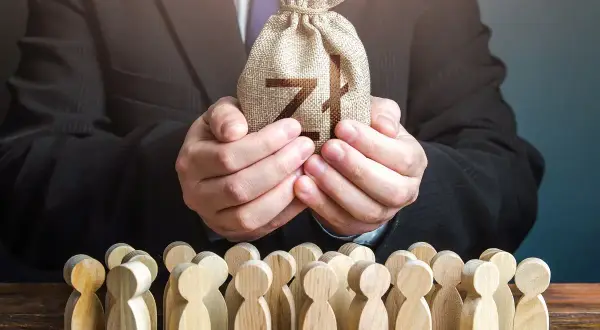 Biznesmen w garniturze trzyma worek z pieniędzmi dla grupy drewnianych ludzików. To symbol, jak siła wyższa może wpływać na losy wielu.
