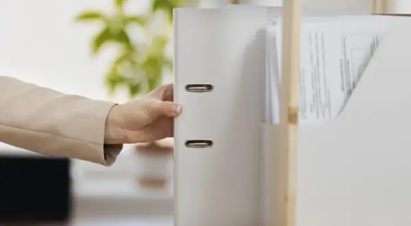 Dłoń w beżowej marynarce sięga po biały segregator z dokumentami. W tle zielone liście, nip-8.