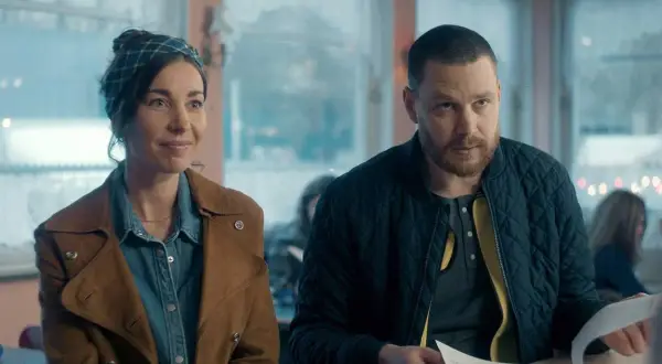 En kvinna med scarf och en man med skägg sitter vid ett bord. Scenen är från en dansk serie på SVT.