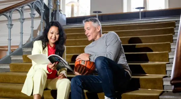 En kvinna och en man sitter på en trappa. Hon läser en tidning, han håller en basketboll. Kanske funderar de på att byta jobb eller inte.