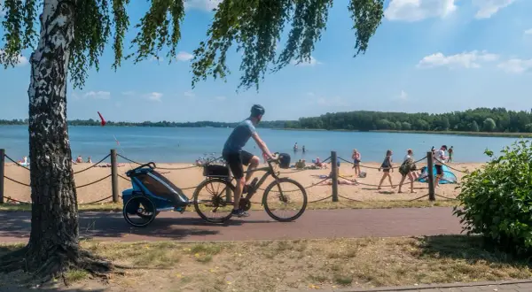 Mężczyzna na rowerze z przyczepką jedzie ścieżką rowerową nad jeziorem. Na plaży opalają się ludzie.