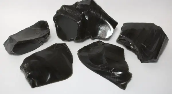 Fünf scharfkantige Stücke glänzenden, schwarzen Obsidiansteins liegen auf weißem Untergrund.