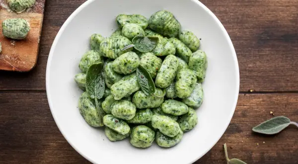 Gnocchi di spinaci freschi, conditi con foglie di salvia, in una ciotola bianca su un tavolo di legno scuro.
