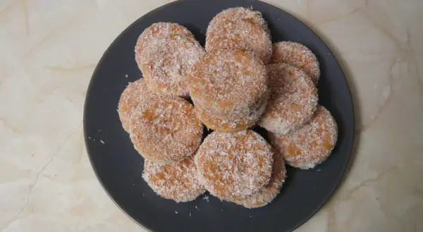 Montón de galletas de coco, doradas y cubiertas de coco rallado, apiladas en un plato oscuro.
