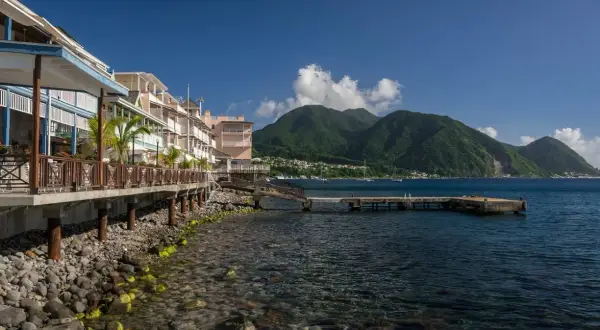 Bâtiments colorés en bord de mer sur l'île Dominique, avec des montagnes verdoyantes en arrière-plan et un quai s'avançant dans l'eau bleue.