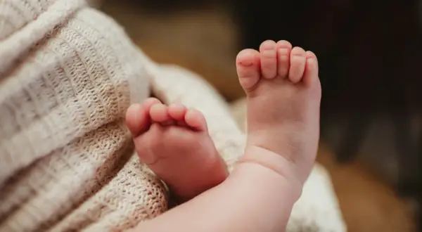 Delikatne, różowe stópki niemowlaka, z widocznymi podwiniętymi palcami u nóg, spoczywają na miękkim, beżowym kocyku.