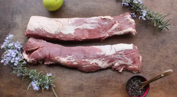 Deux pièces de filet mignon cru sur une planche en bois, accompagnées de romarin fleuri, d'une lime et de baies poivrées.
