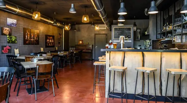 Interno di un locale con tavoli, sedie, un bancone in legno grezzo e sgabelli. Sul bancone due birre, pronte per essere gustate dal baldo birraio.