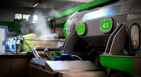 Macchinari industriali in una fabbrica, con tubi e vapore. Si intravede un cartello con "53", forse legato alla produzione di olio di sansa cos'è.