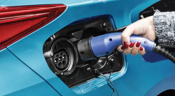 Dłoń z czerwonym lakierem na paznokciach wkłada niebieską wtyczkę do gniazda ładowania samochodu. To przyszłość motoryzacji – hybryda plug-in gotowa do jazdy.