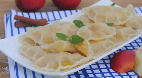 Pyszne pierogi z jabłkami, posypane cynamonem, z listkami mięty i kawałkami jabłek w tle.