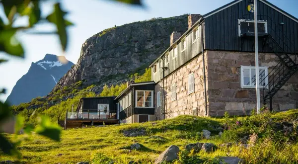 STF Kebnekaise fjällstation badar i solljus. En stenbyggnad med mörka trädetaljer reser sig mot en dramatisk bergsklippa.
