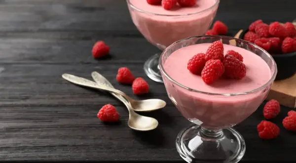 Två glas med hallonmousse, toppade med färska hallon. Två skedar ligger bredvid på ett mörkt träbord.