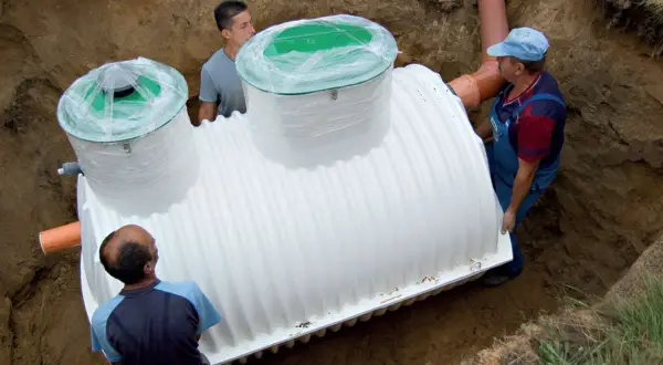 Trzej mężczyźni montują białe szambo betonowe czy plastikowe w wykopie.