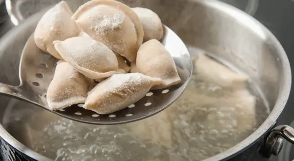 Gotowanie pierogów: na łyżce cedzakowej świeżo uformowane pierogi czekają na wrzucenie do wrzątku. W garnku bulgocze woda, gotowe do przyjęcia pysznego farszu.