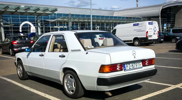 Biały Mercedes W190 z lat 80. parkuje przed nowoczesnym budynkiem lotniska. Klasyk w świetnym stanie.