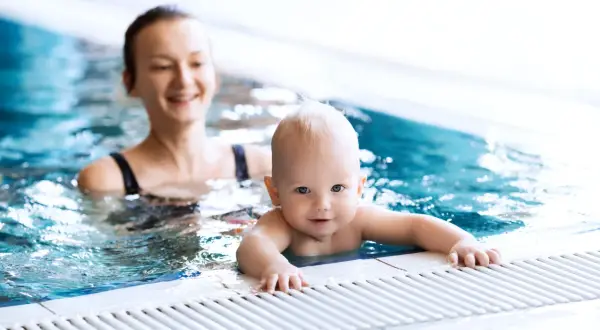 Uśmiechnięty niemowlak w basenie, trzymający się brzegu. Mama w tle obserwuje go z radością.
