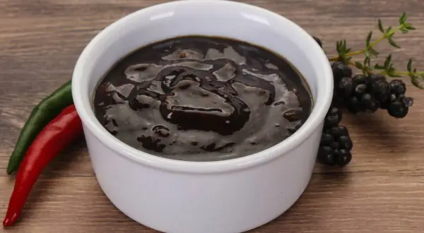Gęsty, ciemny sos pieprzowy w białej miseczce, obok czerwona i zielona papryczka chili oraz gałązka jeżyn.