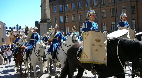 Livgardets dragonmusikkår marscherar i parad. Musiker i blå uniformer spelar på instrument och rider på vita hästar.