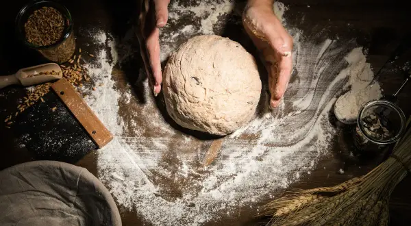 Hände formen einen Teigball auf einer mit Mehl bestäubten Holzoberfläche. Körner, ein Schaber und ein Gärkorb liegen daneben.