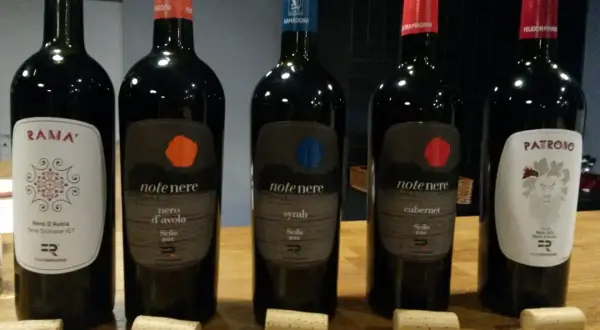 Pięć butelek wina, w tym dwie z etykietą "note nere nero d'avola sicilia doc", stoją na drewnianym stole.