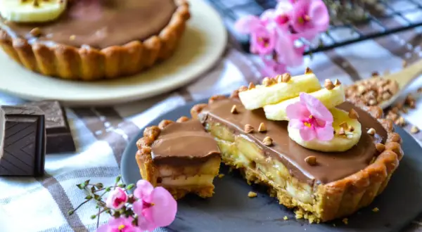 Tarte banane chocolat, garnie de rondelles de banane, de fleurs roses et de noisettes concassées. Un délice gourmand !
