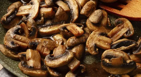 Gebratene Champignons in einer Pfanne, dampfend und glänzend, mit einem hölzernen Kochlöffel.