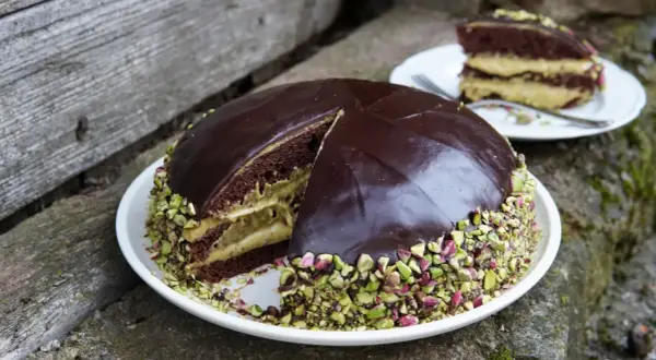 Torta al cioccolato con crema al pistacchio, ricoperta di glassa lucida e granella di pistacchio. Una fetta è già servita.