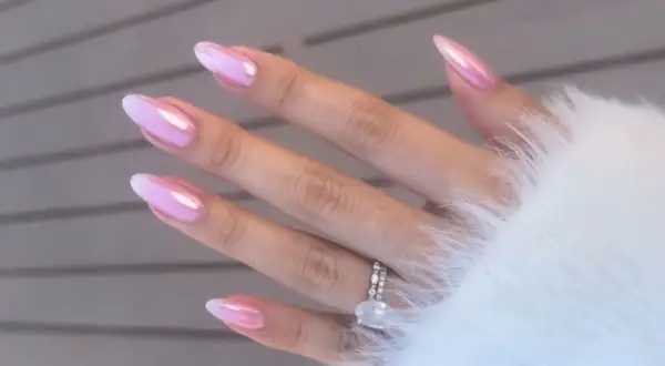 Dłoń z paznokciami w stylu bubble gum, ozdobionymi błyszczącym różowo-białym ombre. Na palcu serdecznym widoczny pierścionek z dużym kamieniem.