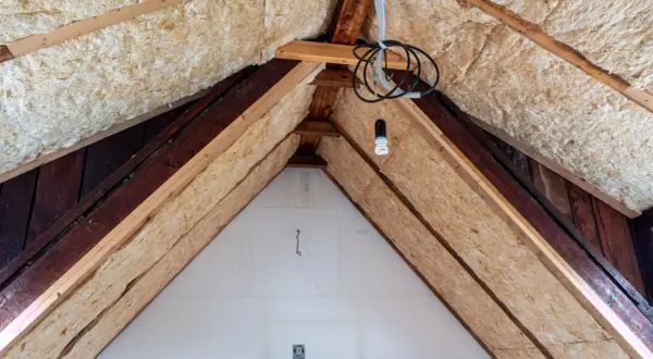 Dämmmaterial wird in einem Dachboden verlegt, um das Haus zu isolieren. Holzbalken und Kabel sind sichtbar.