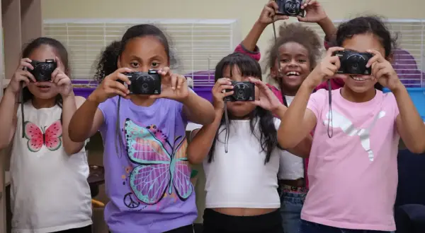 Five girls hold up old cameras, learning what to do with them by taking pictures.
