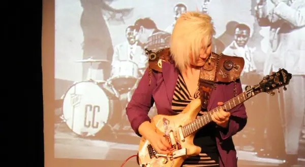 Eine deutsche Gitarristin mit blondem Haar spielt auf einer E-Gitarre vor einer Projektion einer Jazzband.