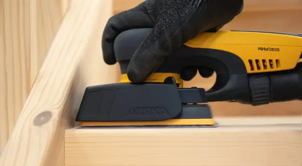Mano guantata utilizza levigatrice elettrica per carteggiare legno grezzo, preparando la superficie per la finitura.