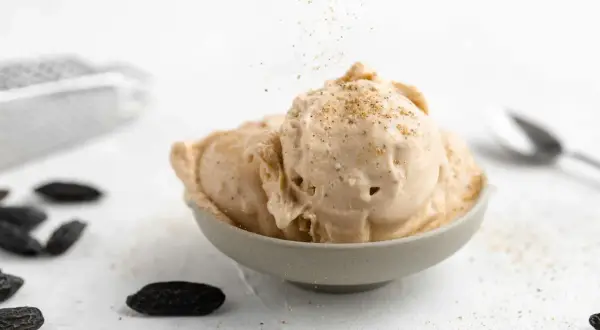 Schüssel mit cremigem Tonka Eis, bestreut mit gemahlenen Tonkabohnen. Dunkle Tonkabohnen liegen verstreut auf dem Tisch.