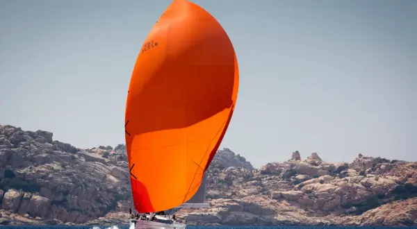 Vela arancione brillante, un gennaker avvolgibile, gonfia al vento su un'imbarcazione a vela che naviga vicino a una costa rocciosa.