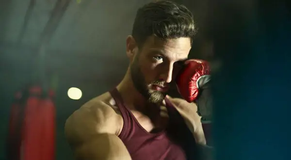 A boxer with a beard and intense blue eyes, wearing a red tank top and a red boxing glove, poses in a gym. He's ready to jump into action.