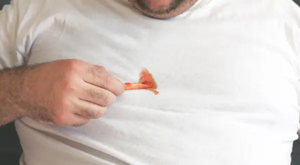 Ein Mann zeigt auf einen Ketchupfleck auf seinem weißen T-Shirt. Solche Fettflecken auf Kleidung sind ärgerlich.