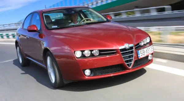Czerwone Alfa Romeo 159 2.4 JTDm pędzi po drodze, kierowca skupiony na trasie.