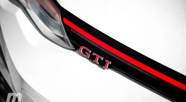 Detalle de un coche blanco con las siglas GTI en rojo y cromo, sobre una franja negra y roja.