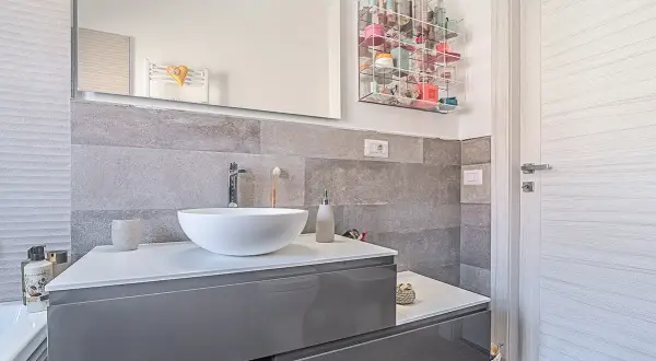 Bagno moderno grigio e legno con lavabo bianco, specchio e porta in legno chiaro. Organizzatore trasparente pieno di cosmetici.