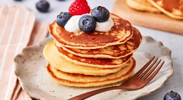 Ein Stapel goldbrauner Pancakes mit Joghurt, Blaubeeren und einer Himbeere obendrauf. Ein köstlicher pancake mit joghurt für ein perfektes Frühstück.
