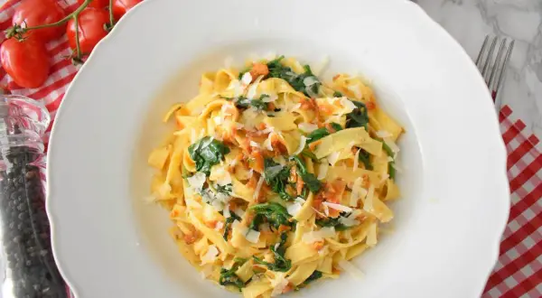 Pyszny makaron ze szpinakiem i pomidorami, posypany parmezanem. Obok świeże pomidorki i młynek do pieprzu.